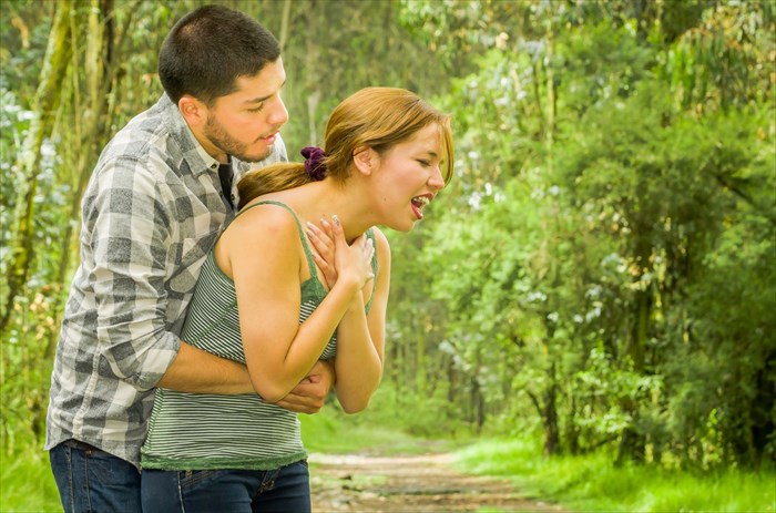 Young woman choking with a man standing behind her, performing the Heimlich maneuver.
